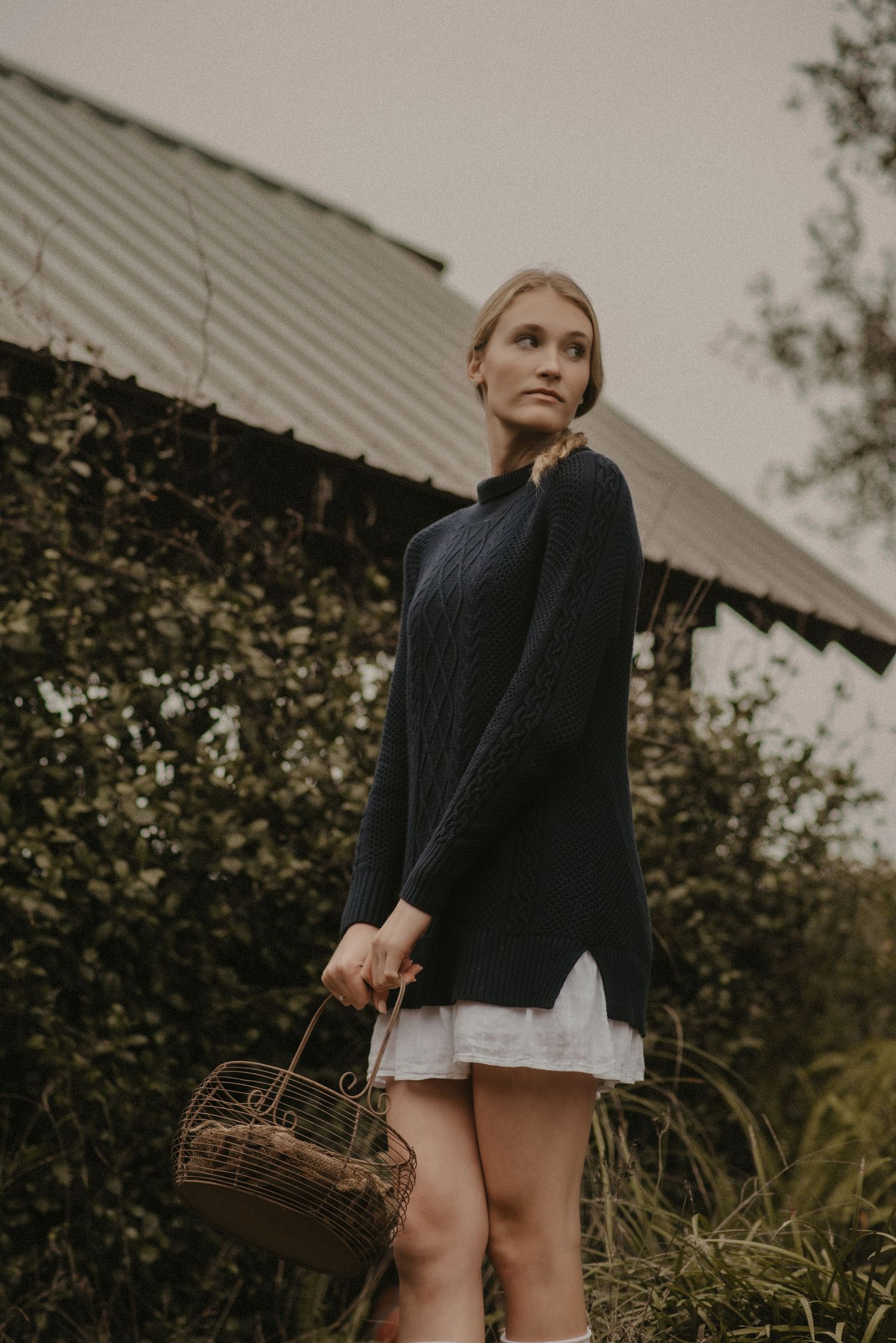 Moody autumn fashion portrait with dark sweater, white dress and woven basket against rustic barn backdrop.