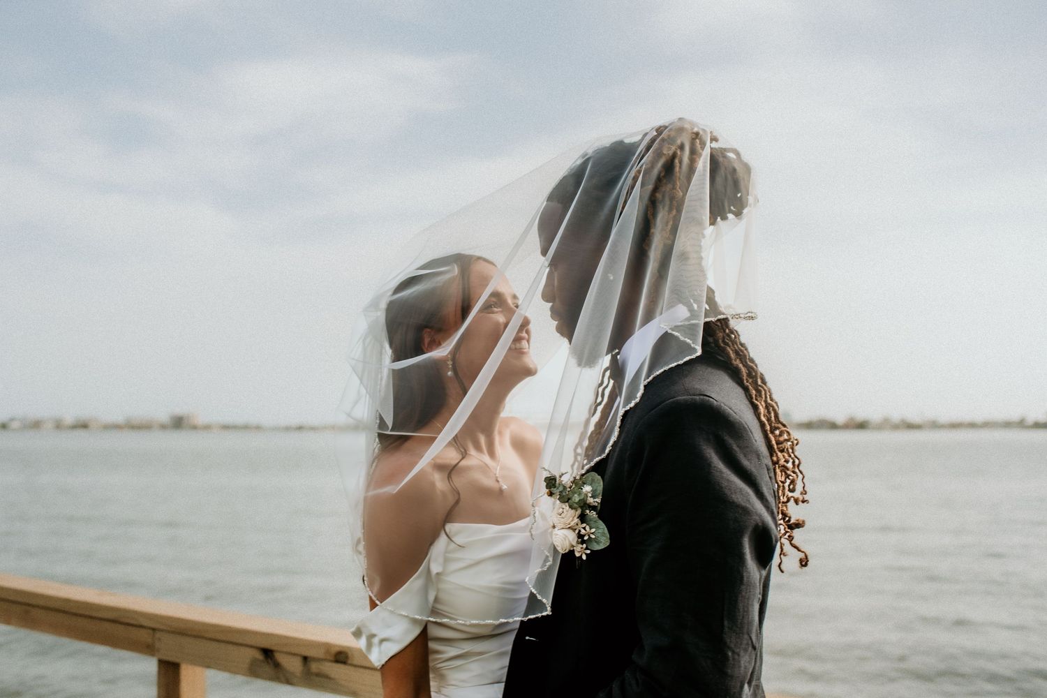 A couple in wedding attire shares an intimate moment on a dock by the water.