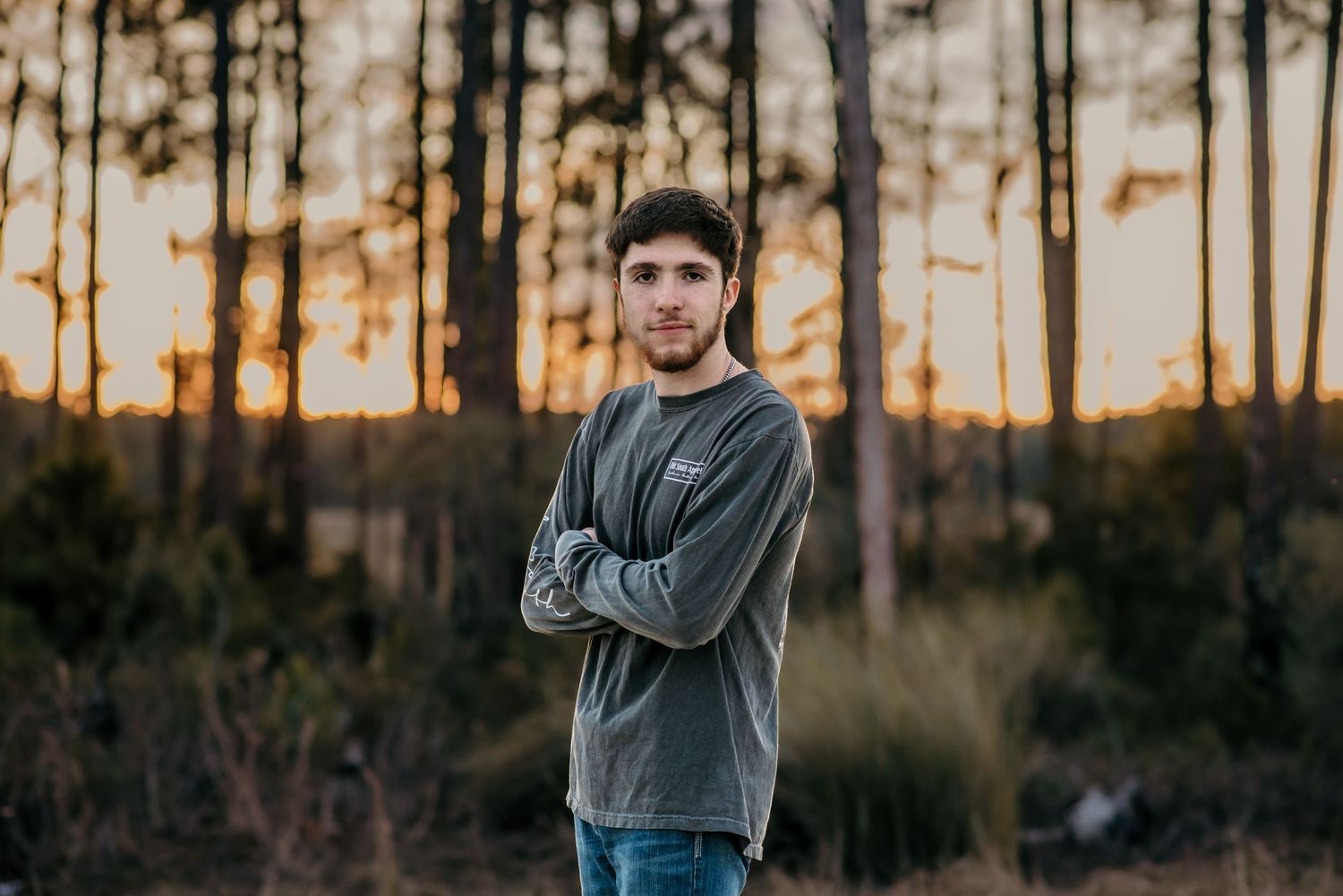 A casual portrait of someone in a grey sweatshirt standing in front of autumn trees at sunset.
