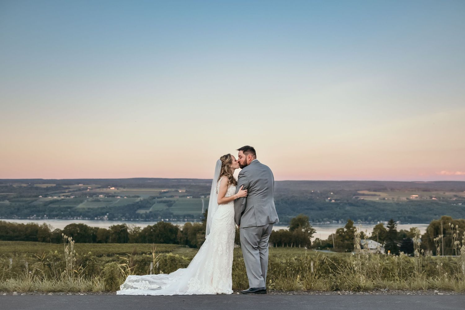 A couple in wedding attire embraces on a hilltop at sunset with a lake view.