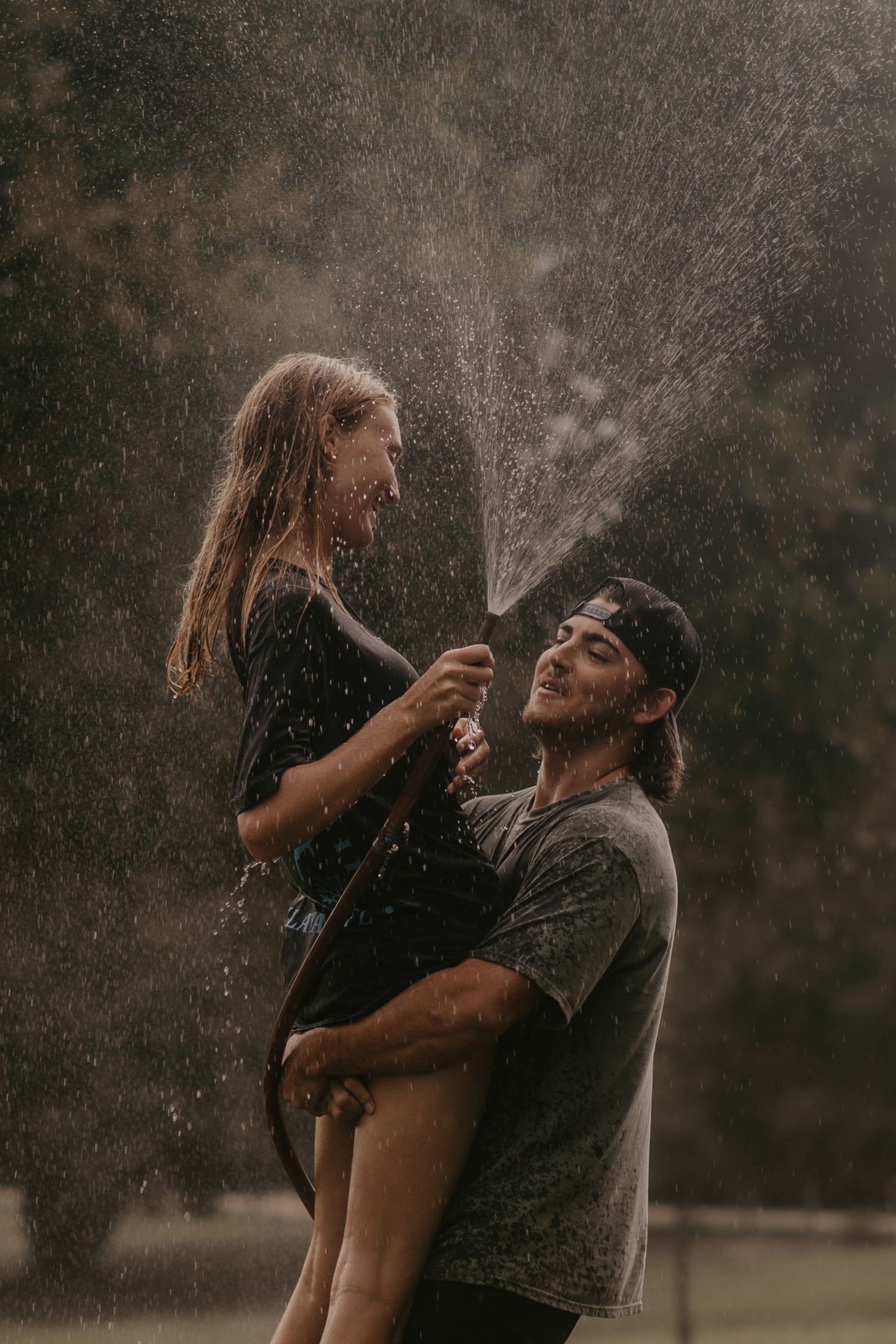 A couple plays in the rain as water sprays between them creating a magical moment in moody lighting.