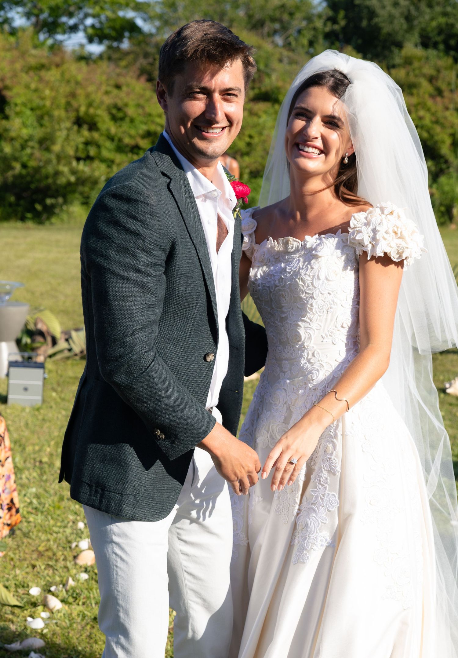 Smiling bride in floral lace gown and groom in dark blazer pose together at outdoor wedding.