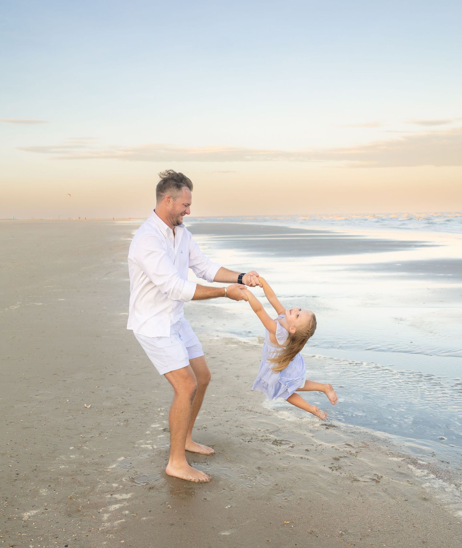 A father joyfully swings his young daughter on a serene beach at sunset, both dressed in white.