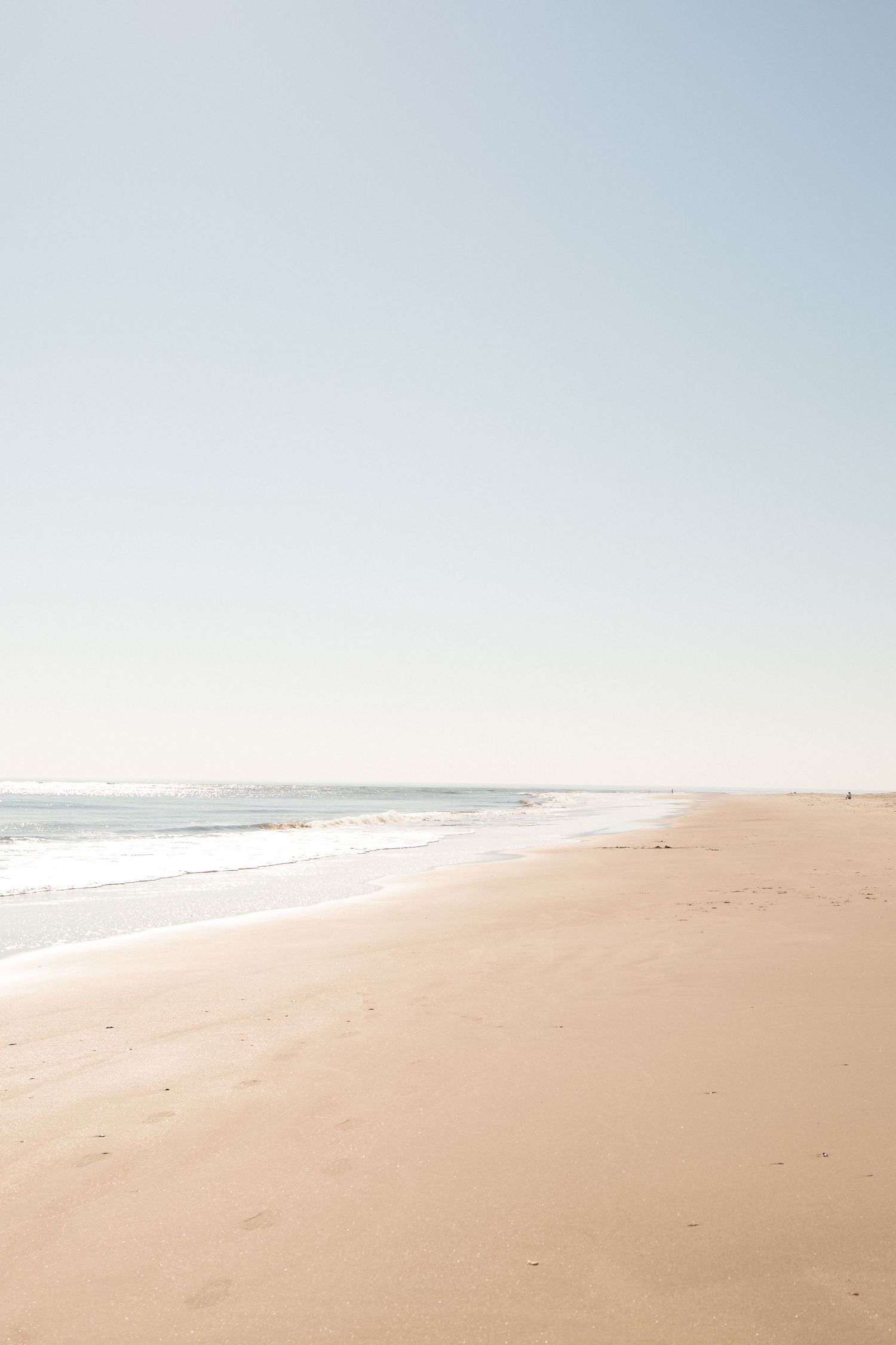 Serene sandy beach stretches along the coastline with gentle waves and clear blue sky on a sunny day.