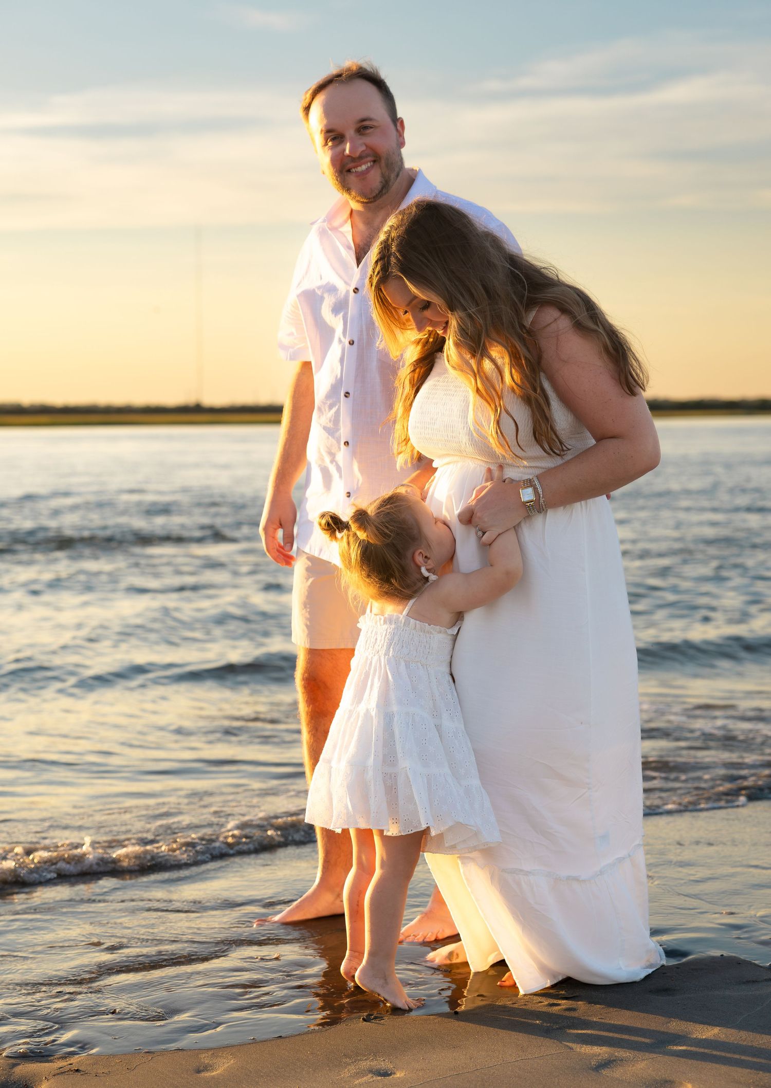 Family of three in white outfits posing on a sandy beach at golden sunset, with waves gently lapping the shore.