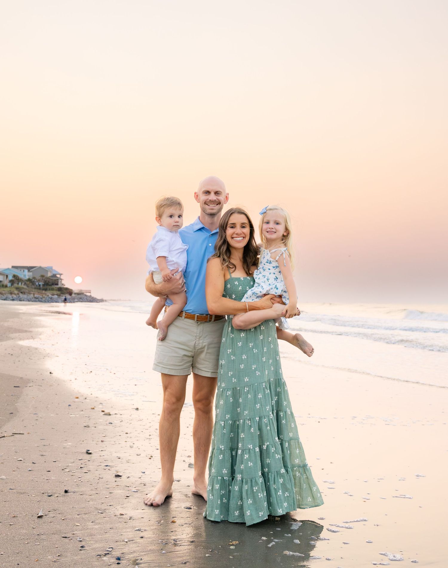 Family of four posing on a beach at sunset, parents holding two young children, wearing pastel summer outfits.