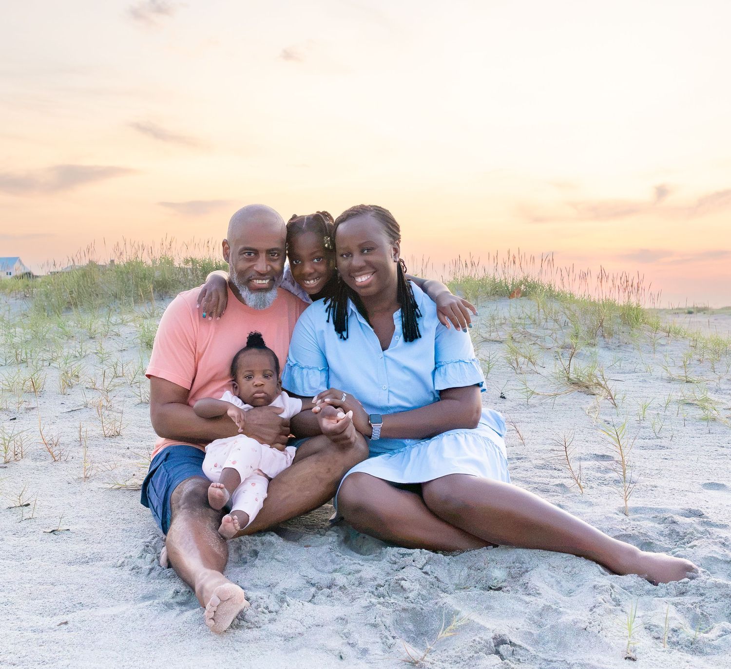 Family of three sitting on beach at sunset, parents smiling with baby on lap, sand dunes and colorful sky behind them.