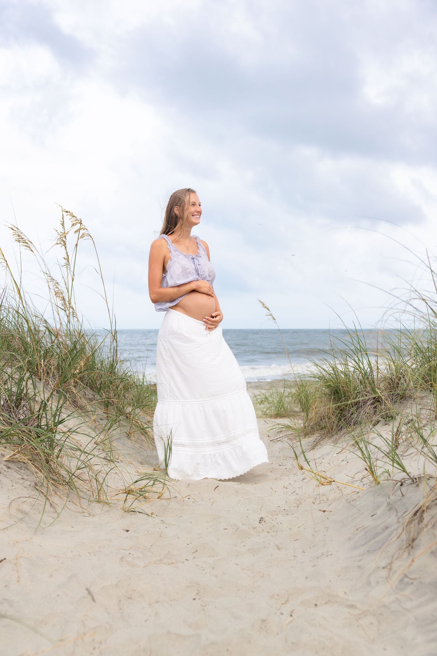 Smiling pregnant woman in white skirt holds baby bump on sandy beach dunes with sea oats and ocean in background.