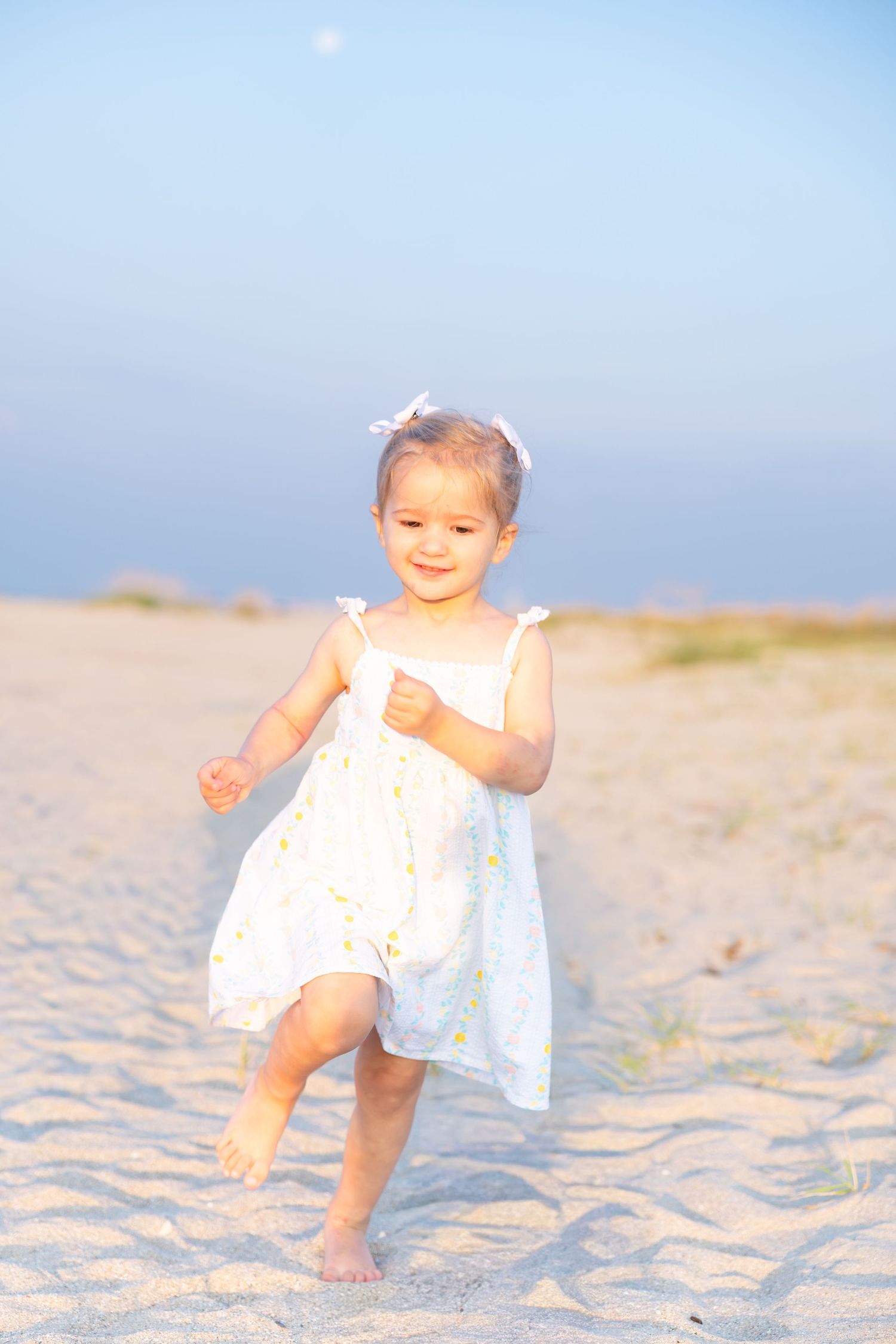 A happy toddler girl in a white dress runs barefoot on a sandy beach under a clear blue sky.