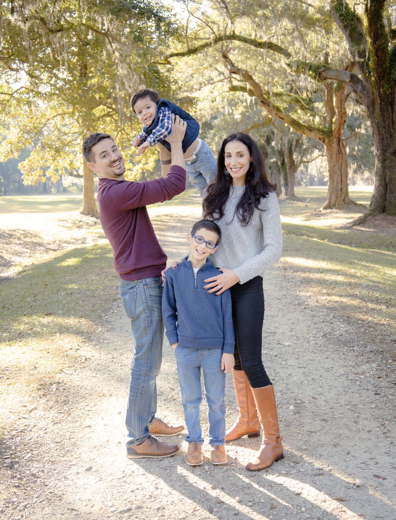 Family of four posing outdoors on a tree-lined path, father lifting toddler while mother and son smile together.