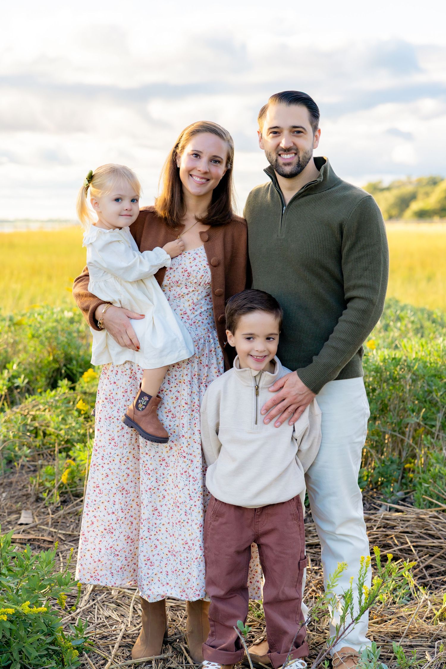 Smiling family of four with a toddler girl, a young boy, and parents posing together in a golden field at sunset.