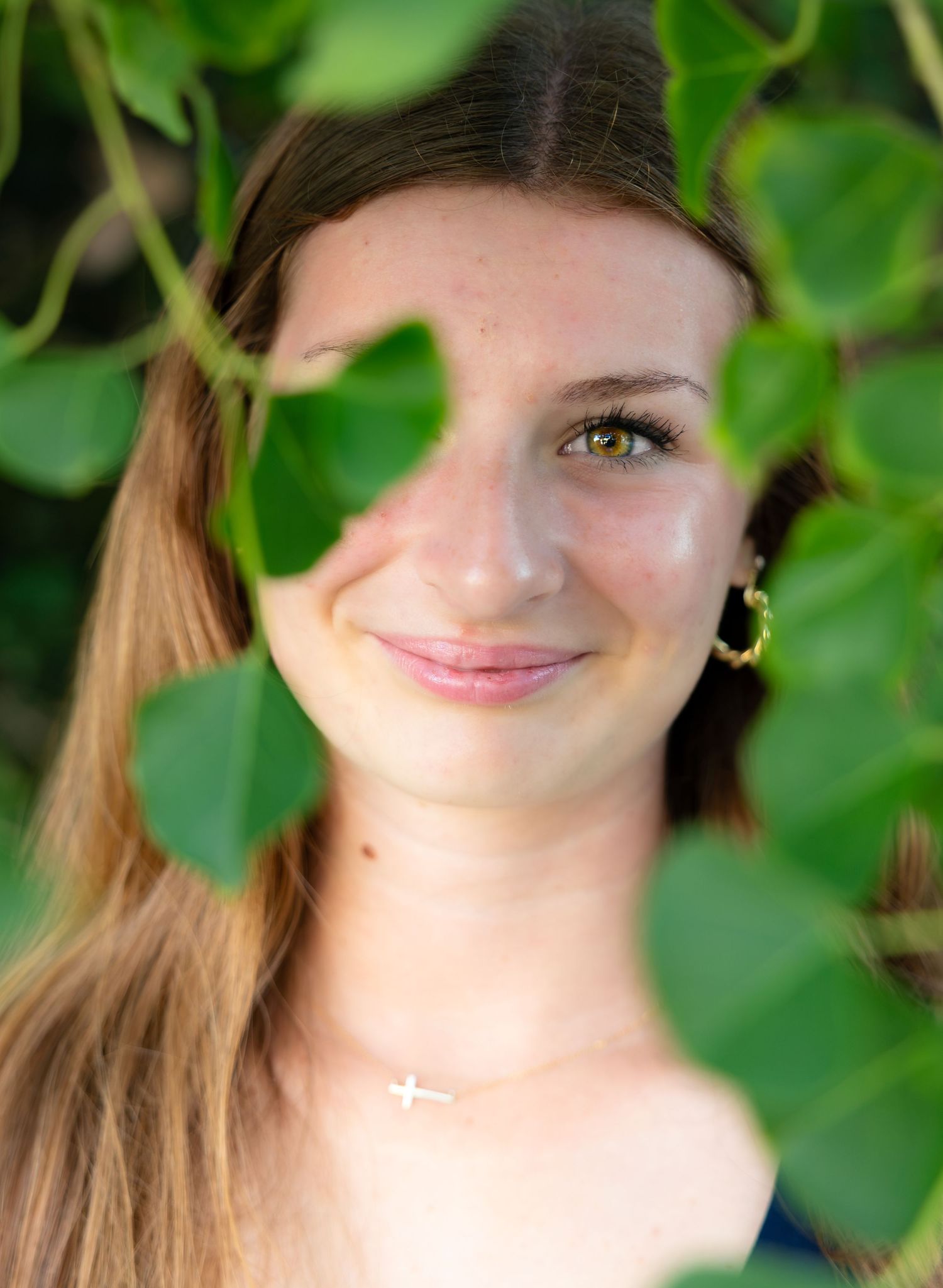 Smiling young woman with long brown hair, gold hoop earring and cross necklace, framed by green leaves in an outdoor portrait.