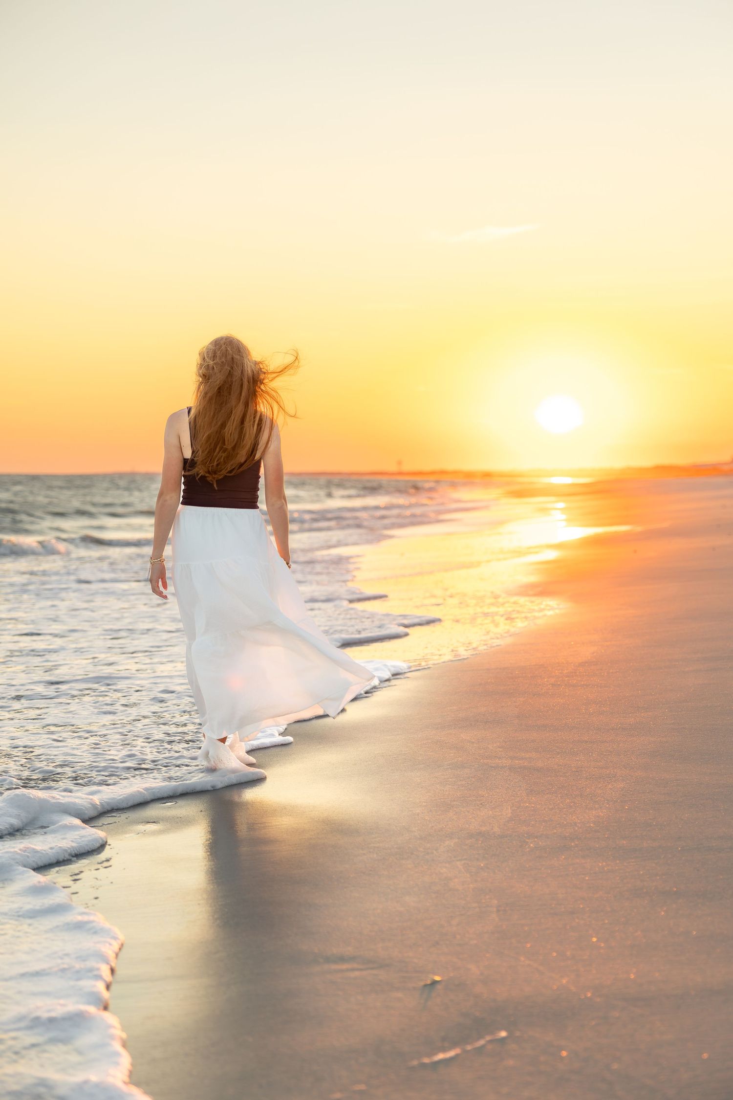 Woman in a flowing white skirt walks barefoot along the shoreline at sunset, with golden light reflecting on the wet sand and gentle waves.