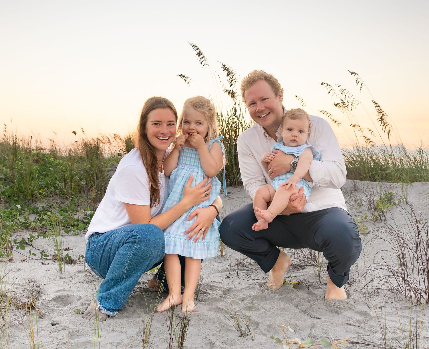 Family of four smiling on sandy beach at sunset with sea oats in background.