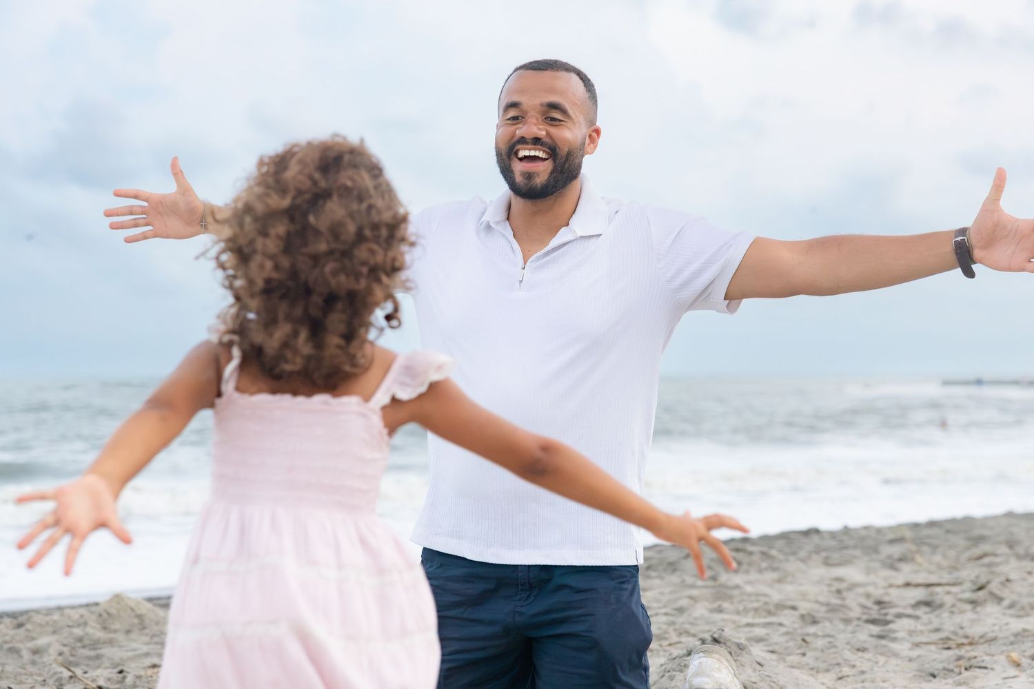A father joyfully opens his arms to embrace his young daughter on a sandy beach by the ocean.
