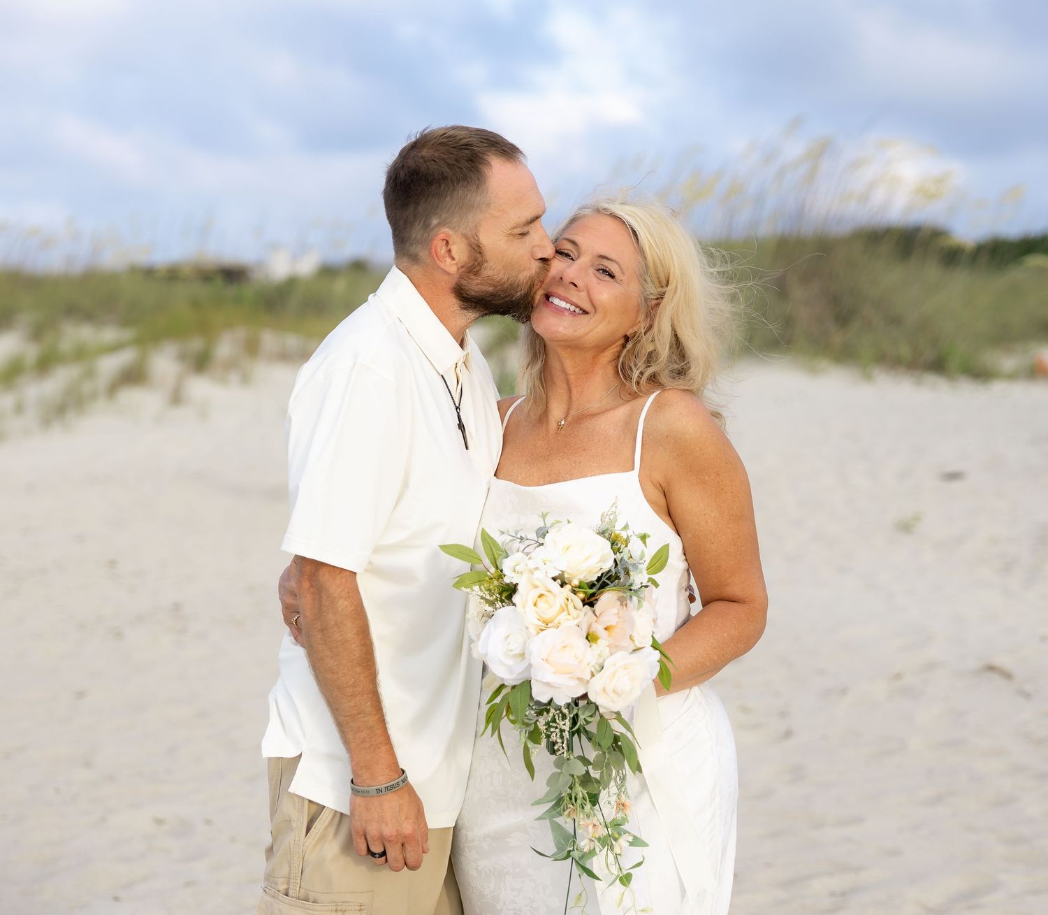 Couple in wedding attire sharing a kiss on a sandy beach, bride holding white floral bouquet.