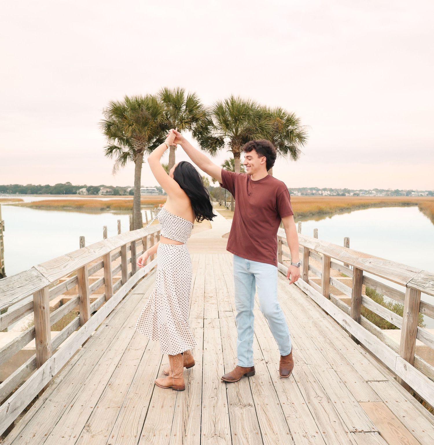 Couple dancing on a wooden pier with palm trees and coastal marsh in the background at sunset.