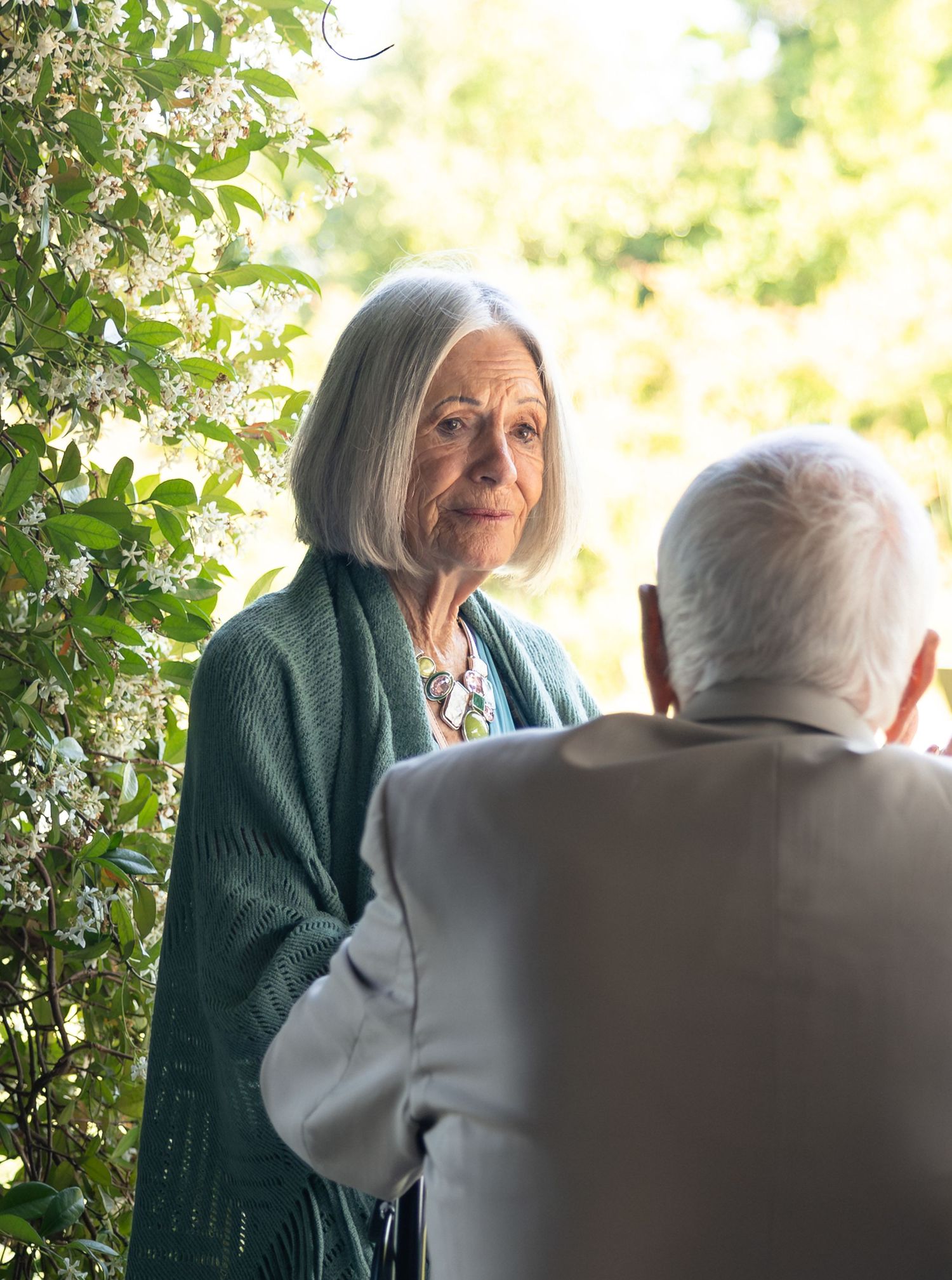 Elderly woman with silver bob hair in green cardigan converses with gray-haired man outdoors near blooming trees.