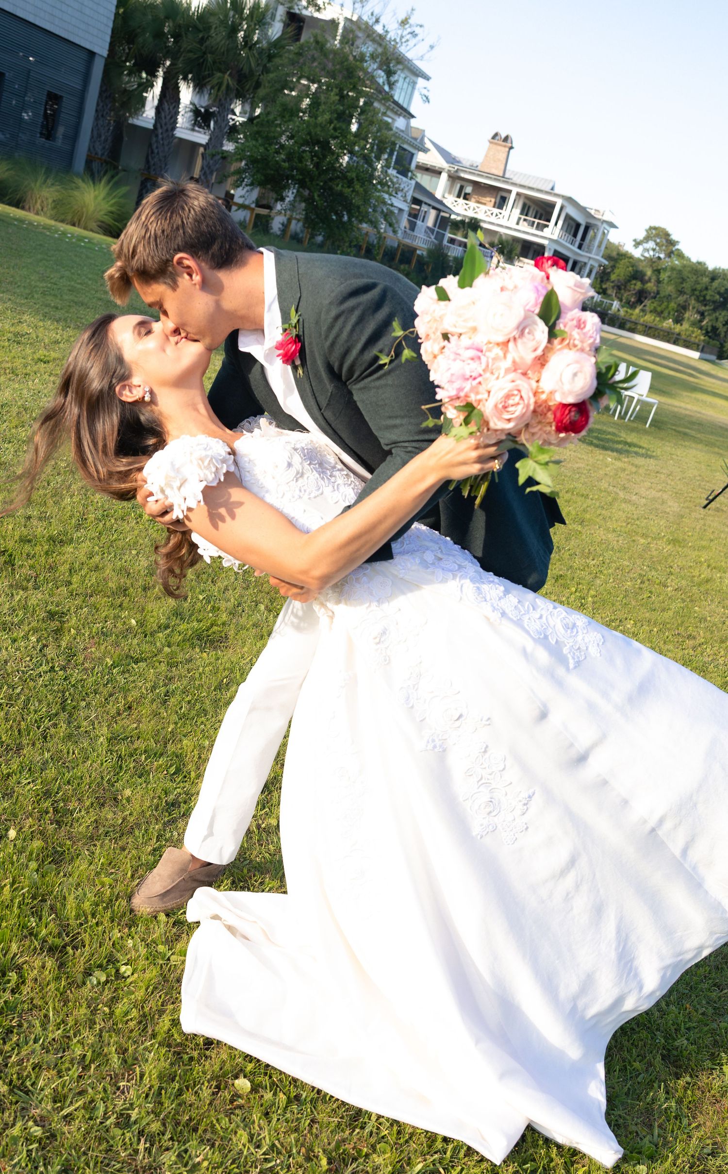 Bride and groom share a romantic dip kiss on green lawn, bride holding pink peony bouquet in white lace wedding gown.