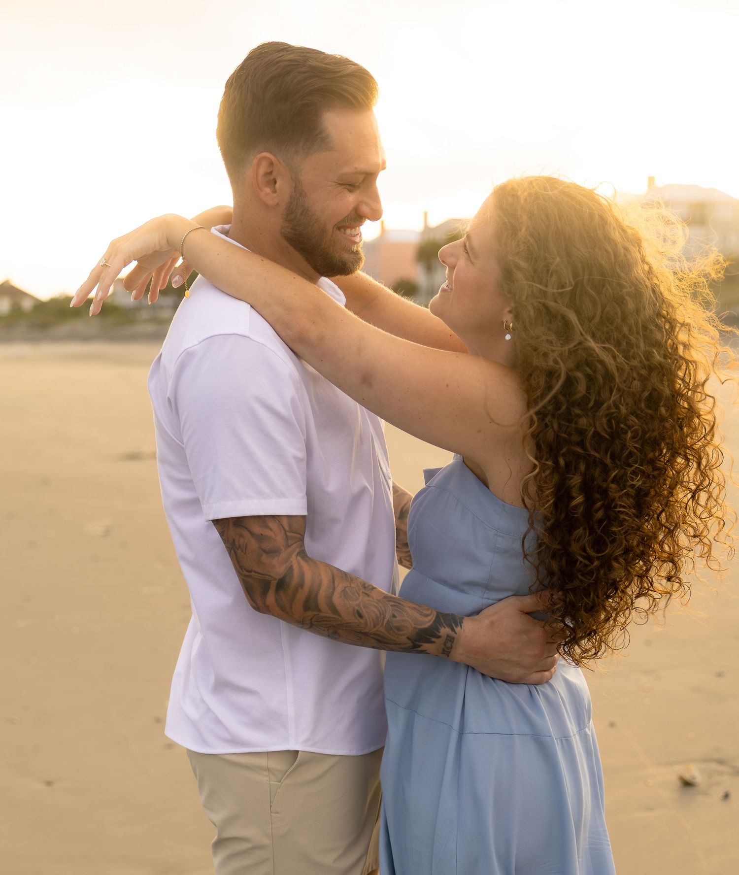Couple embracing on a beach at golden sunset, man with tattoos in white shirt, woman in blue dress.