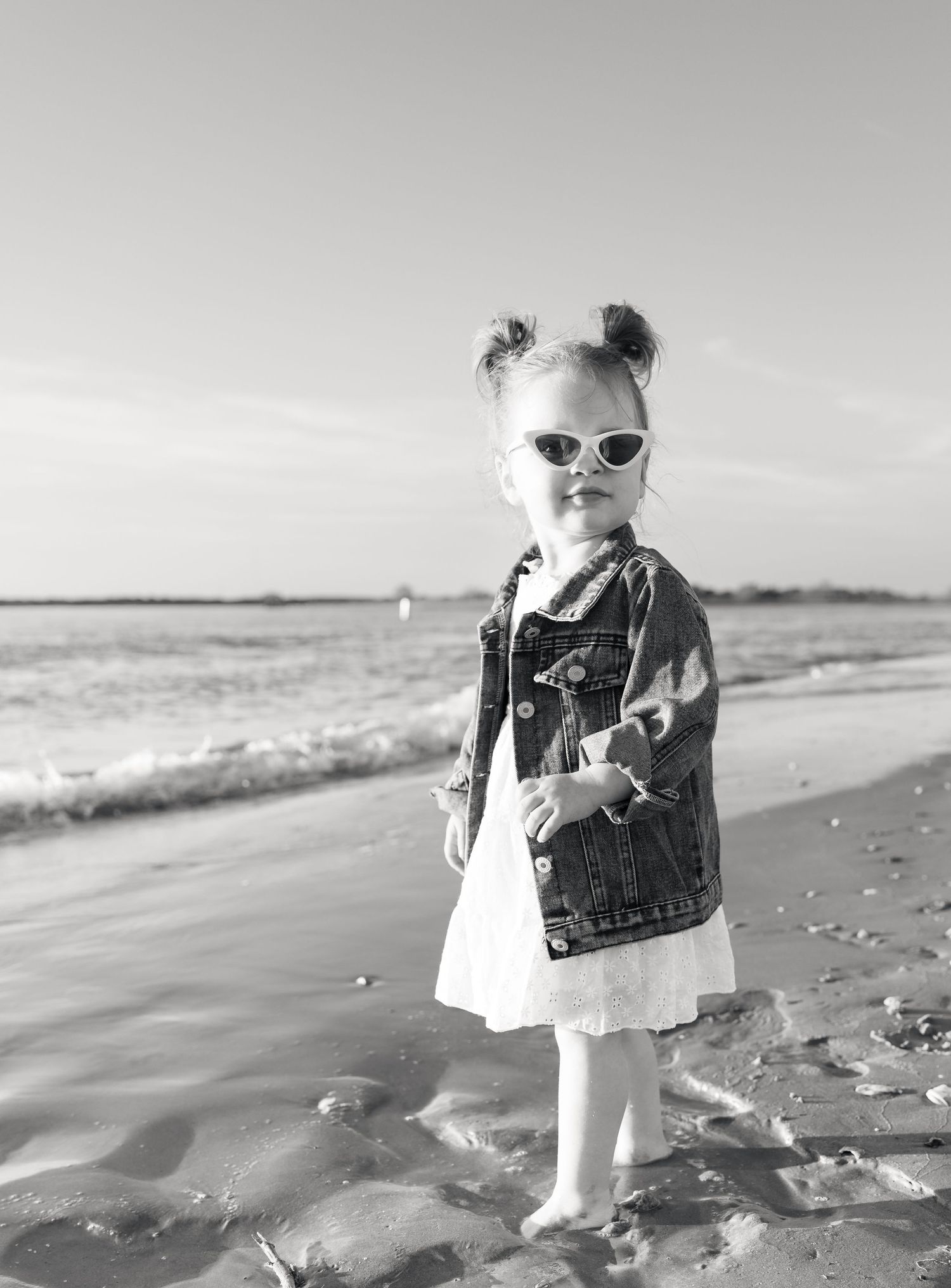Black and white photo of a stylish toddler girl in sunglasses and denim jacket standing on a sandy beach.