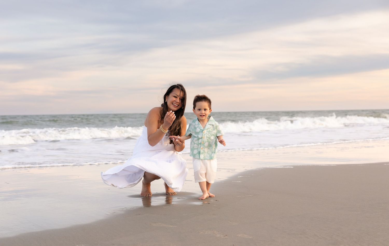 Mother in white dress laughing with toddler son running along ocean shoreline at sunset.