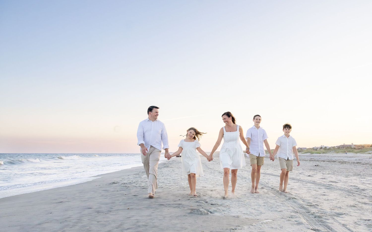 Family of five holding hands walking on a sandy beach at sunset, dressed in coordinating white outfits.