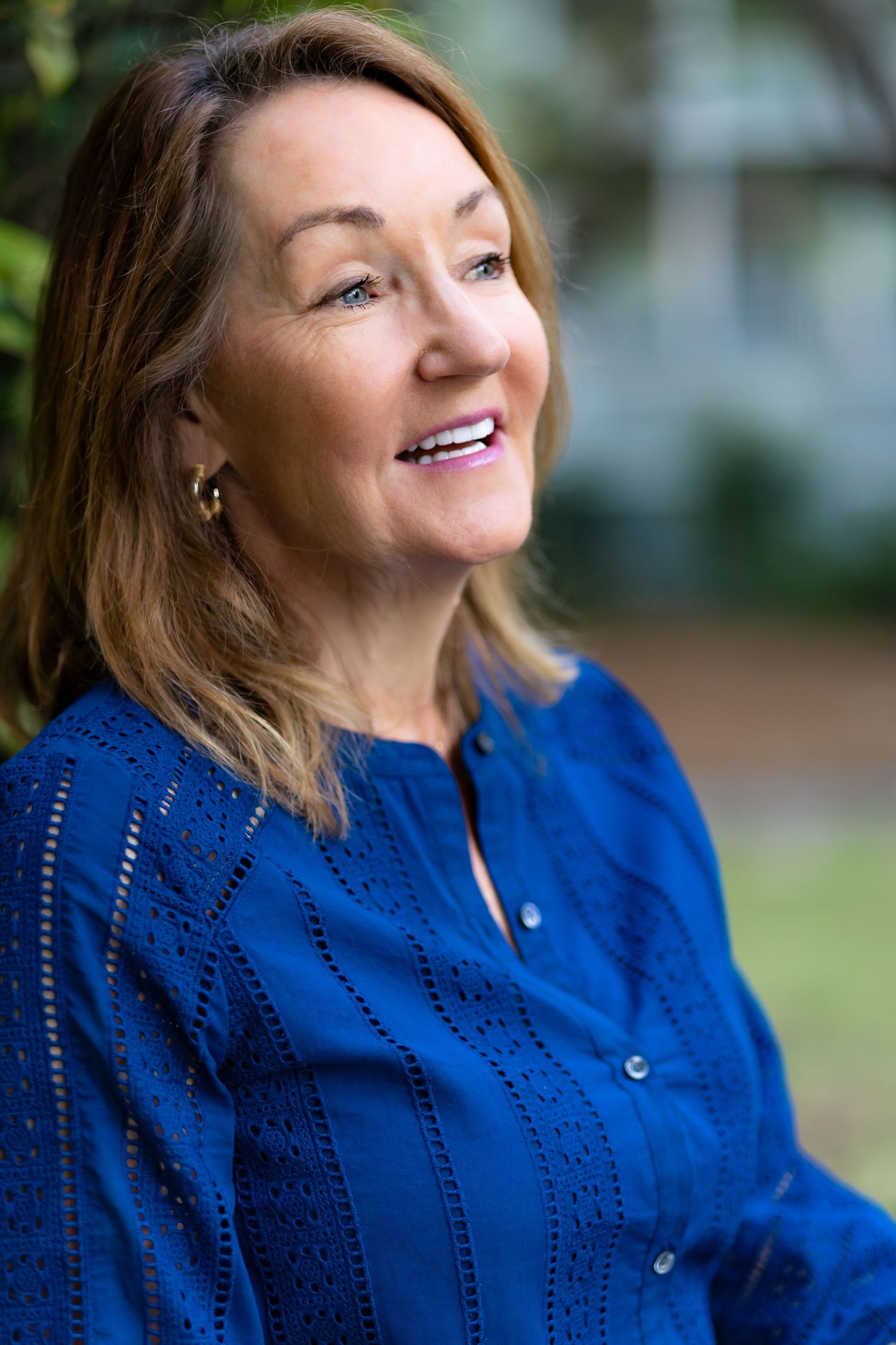 Smiling middle-aged woman with brown hair wearing a blue eyelet blouse outdoors.