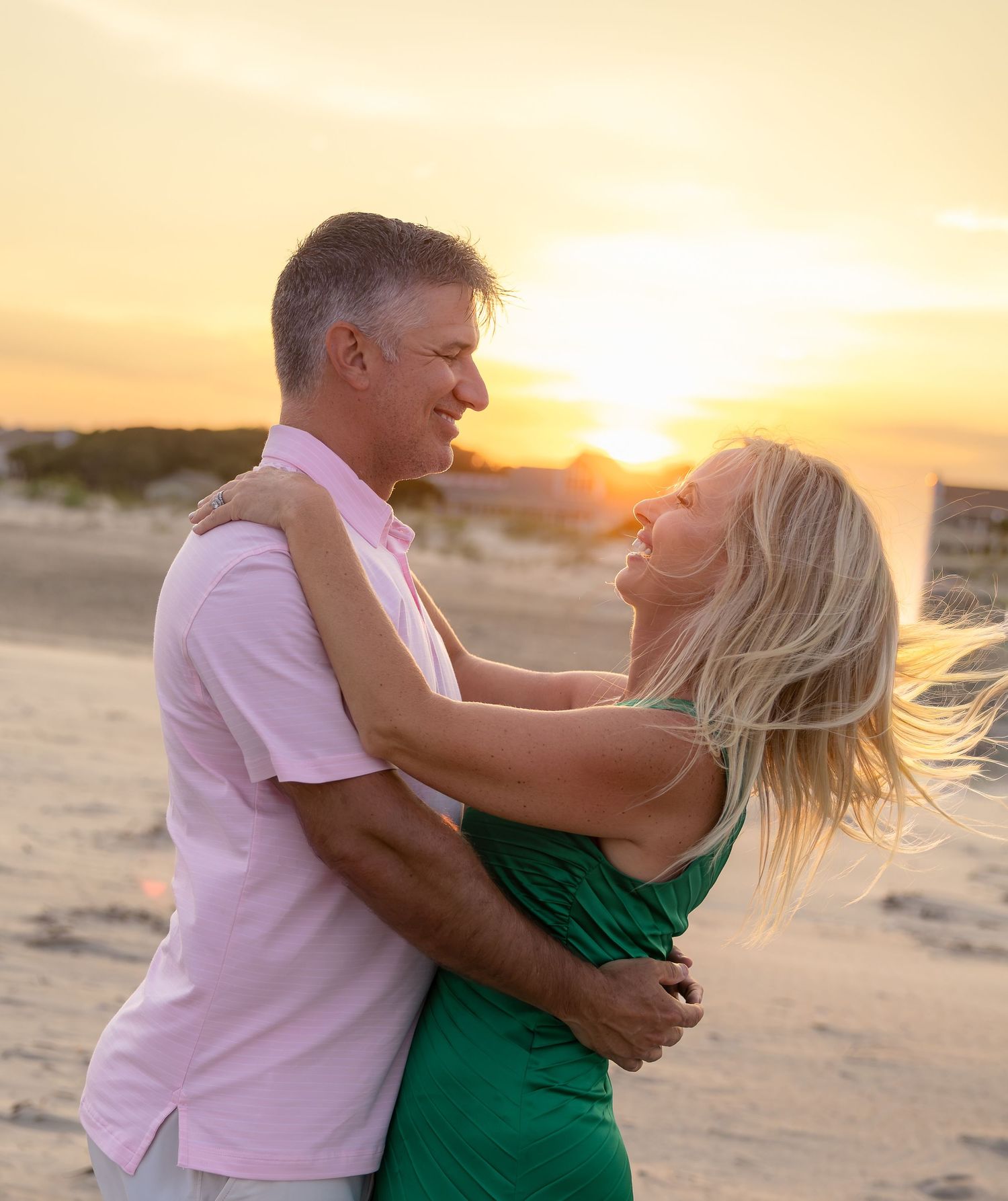Smiling couple embracing and dancing on a sandy beach at golden sunset, the woman in a green dress and the man in a pink shirt.