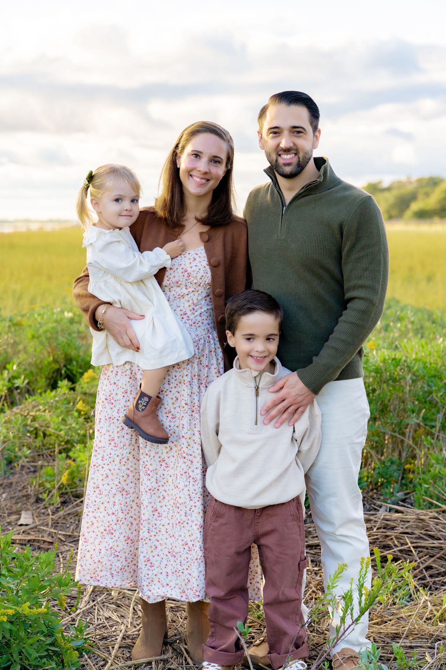 Smiling family of four posing outdoors in a golden grass field, with parents holding a toddler girl and standing beside a young boy.