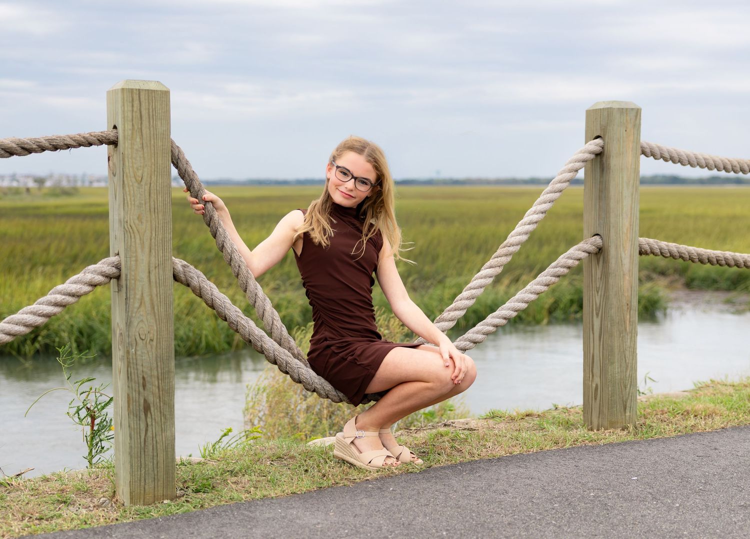 Young woman with glasses in a maroon dress sits on a grassy edge beside a rope fence overlooking a marsh and calm water.