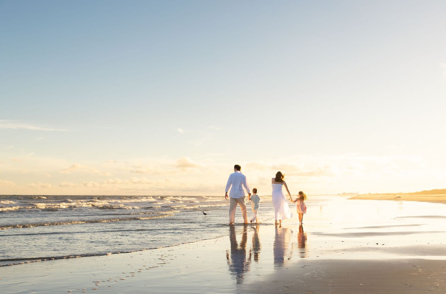 Family in white walking hand-in-hand on a sunny beach at sunset with gentle waves.