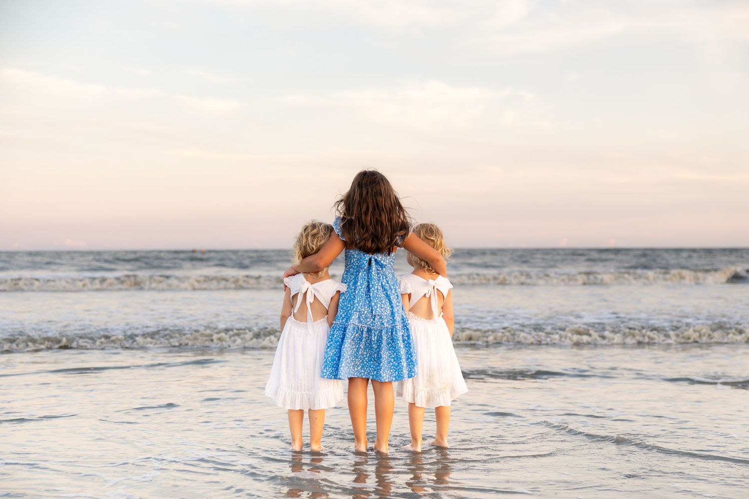 Three girls in white and blue dresses stand together at the ocean's edge, gazing at the sea at sunset.