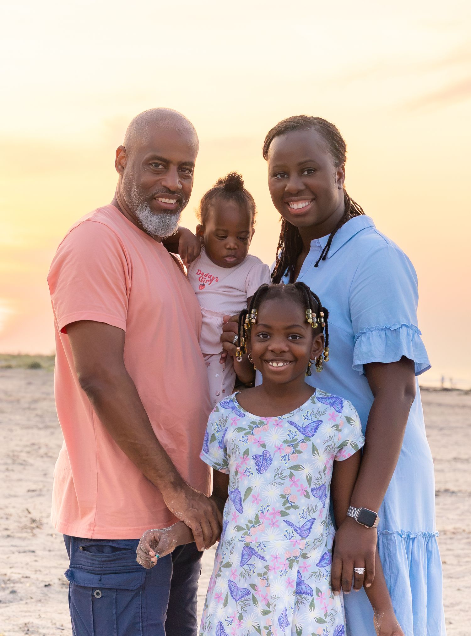 Family of four poses on sandy beach at golden sunset, parents smiling with two young daughters in colorful summer dresses.