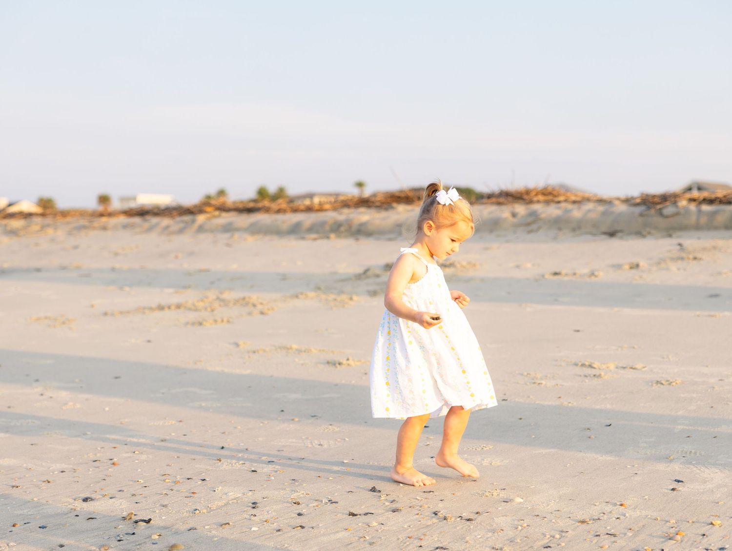 A toddler girl in a white dress walks barefoot on a sandy beach in soft golden light.