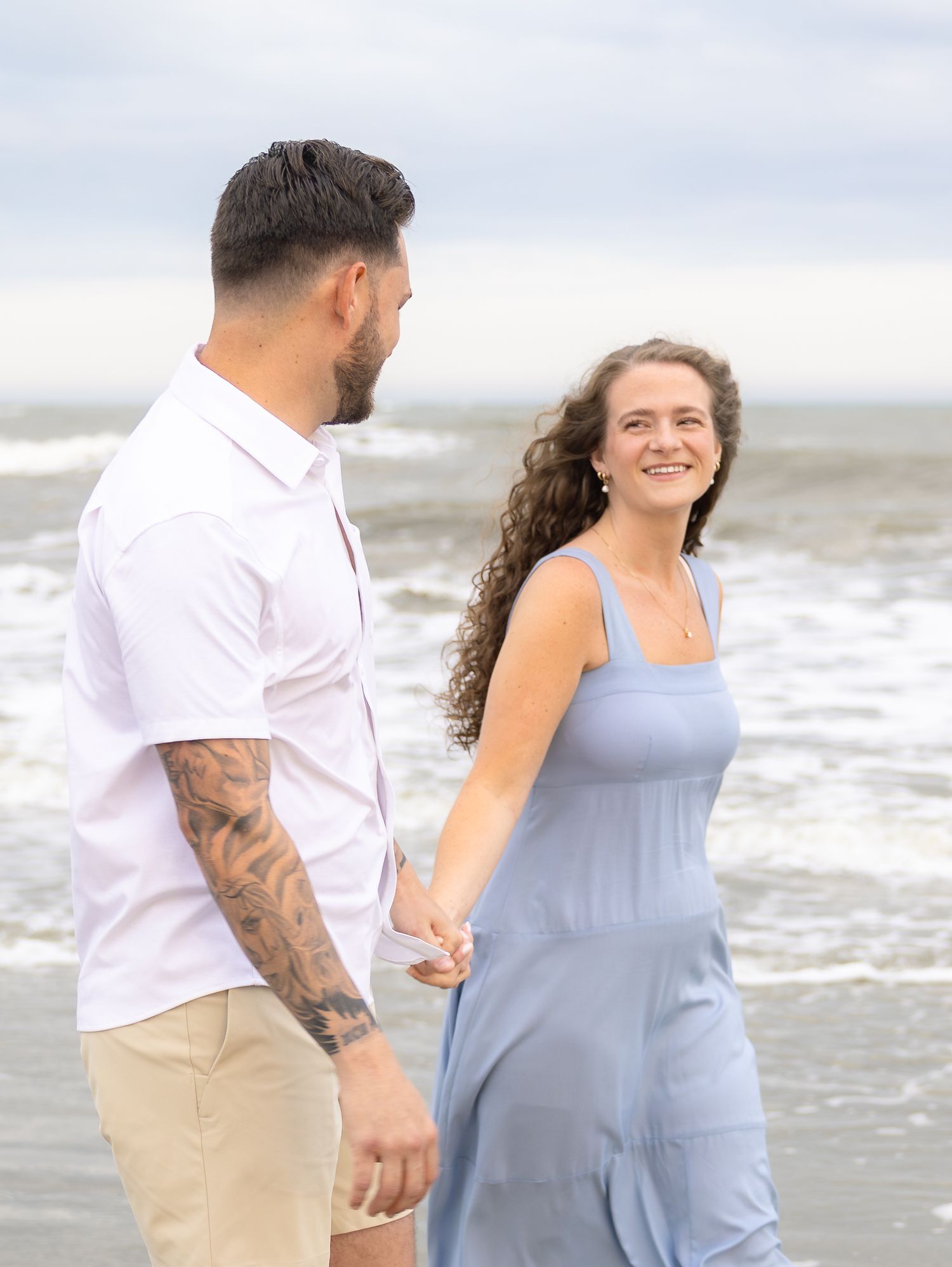 Couple holding hands on a beach, man with tattoos in white shirt, woman in blue dress, ocean waves behind them.
