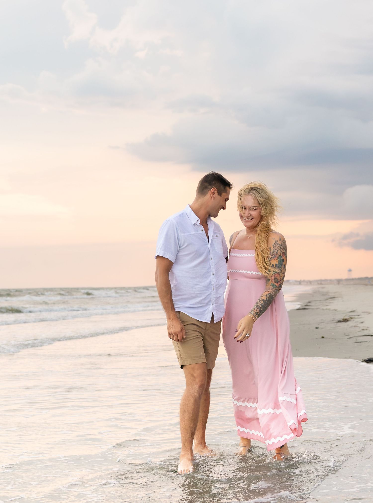 Couple walking barefoot on beach at sunset, man in white shirt and shorts, woman in pink maxi dress with tattoos.