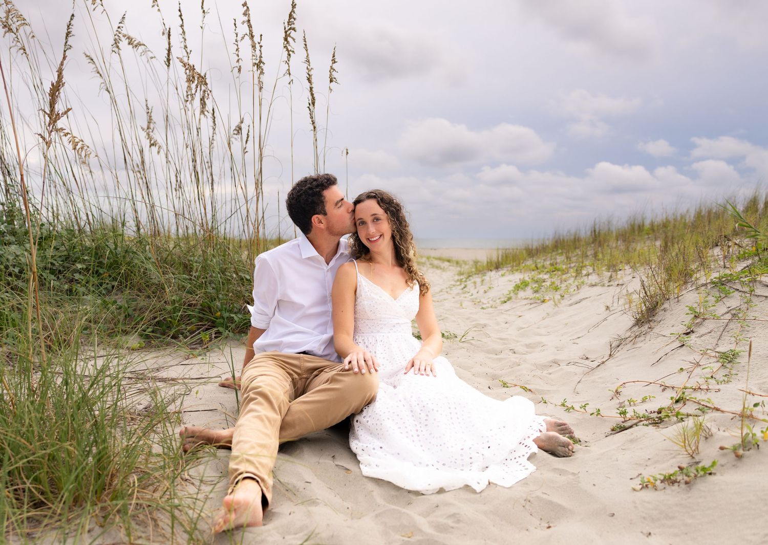 A couple sits on sandy beach dunes, man kissing woman's cheek, she wears white dress, sea oats in background.