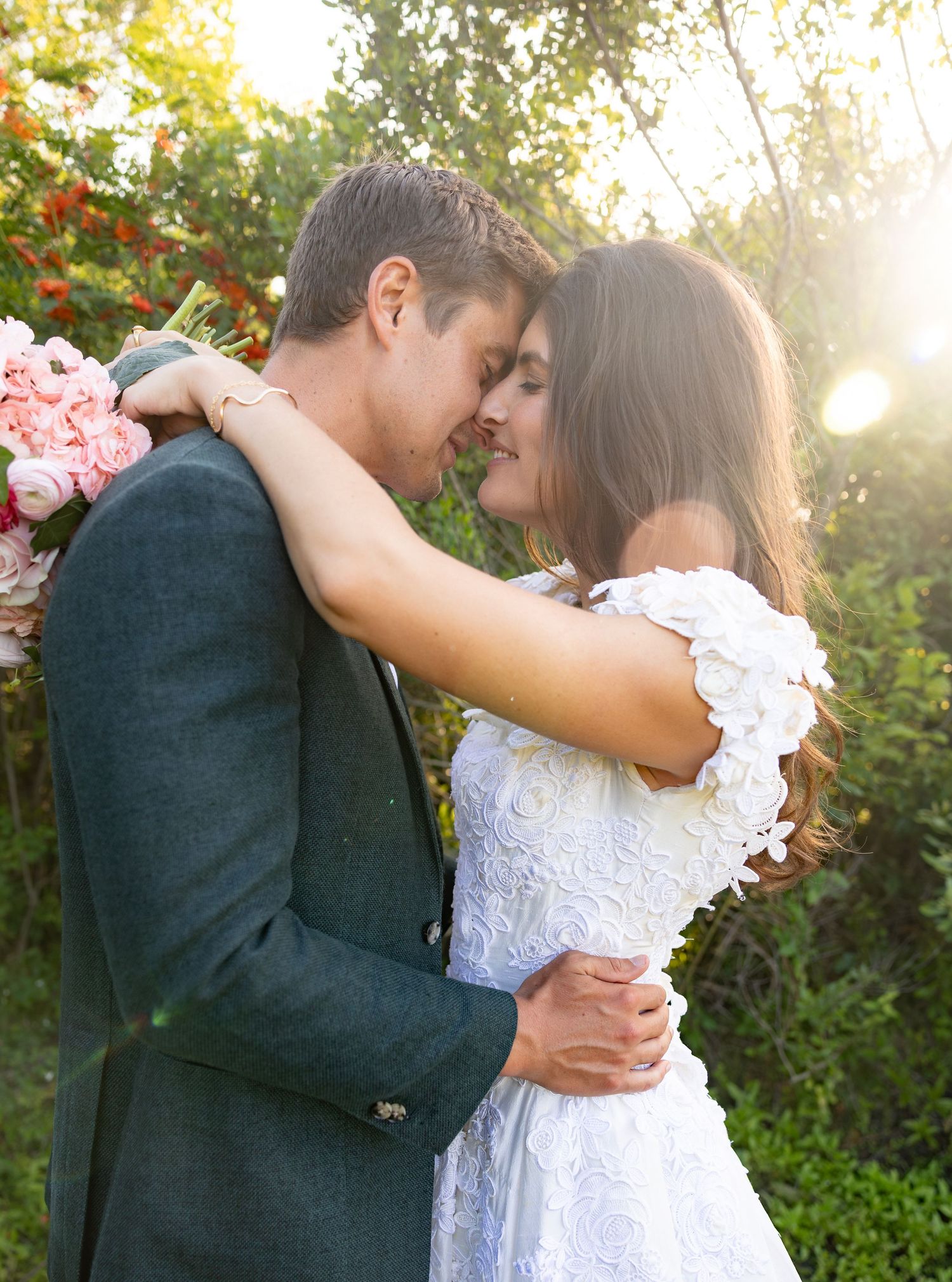 Couple in wedding attire sharing a romantic moment outdoors, groom in dark suit, bride in white lace dress holding pink flowers.