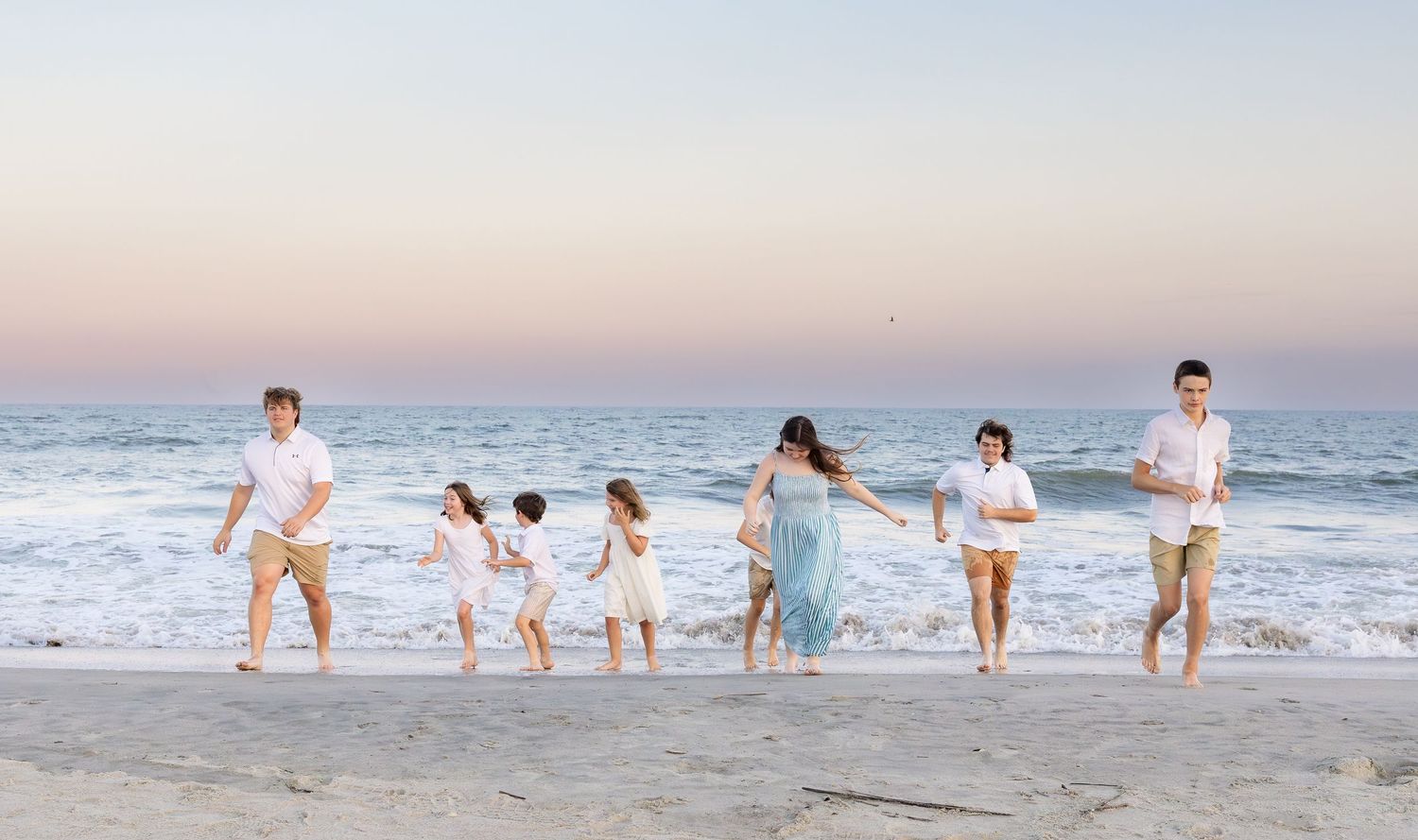 Family holding hands walking along a sandy beach at sunset, wearing white and light blue outfits.