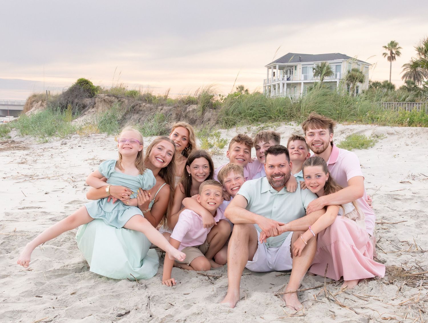 Large family smiling together on a sandy beach at sunset, wearing coordinated pink and mint outfits.