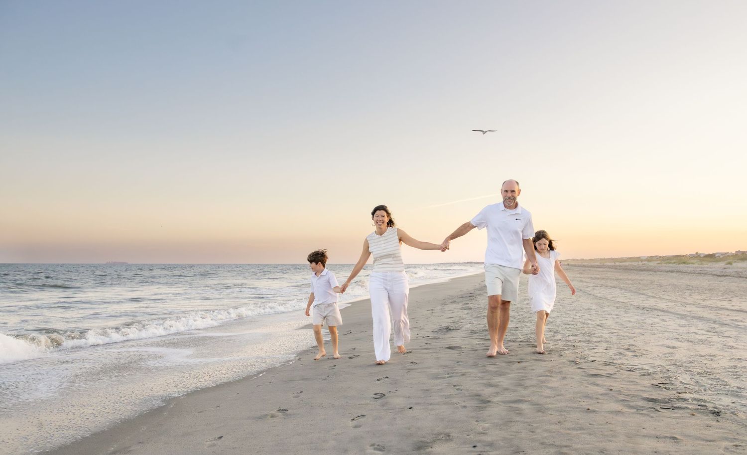 Family in white outfits walking hand-in-hand on a sandy beach at sunset with calm ocean waves nearby.