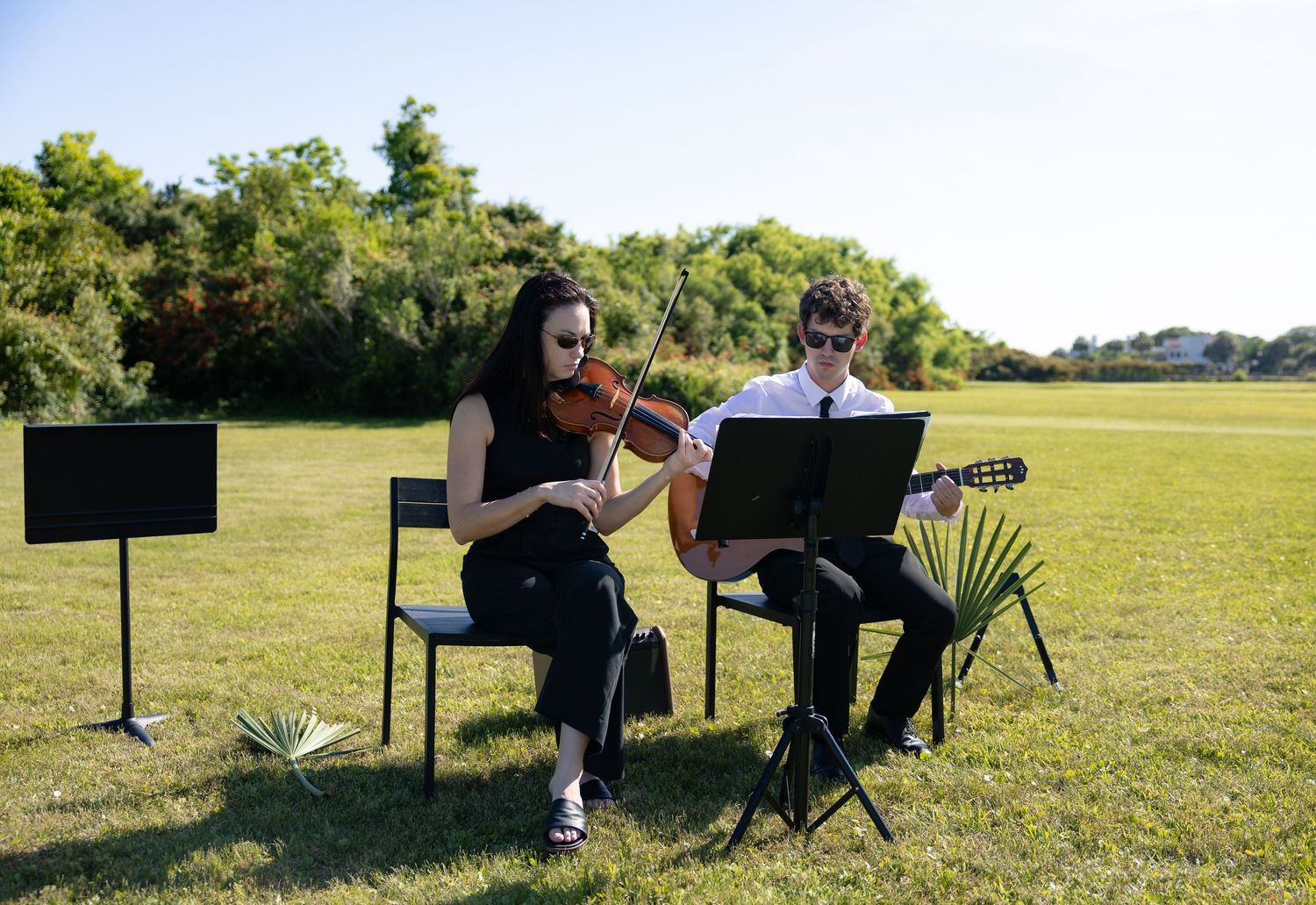 Female violinist and male guitarist perform outdoors on a sunny green lawn.