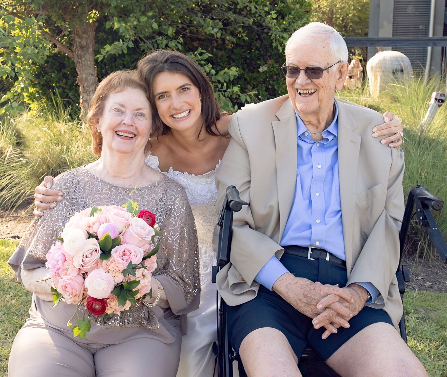 Elderly couple and bride posing together outdoors with pink bouquet at wedding celebration.