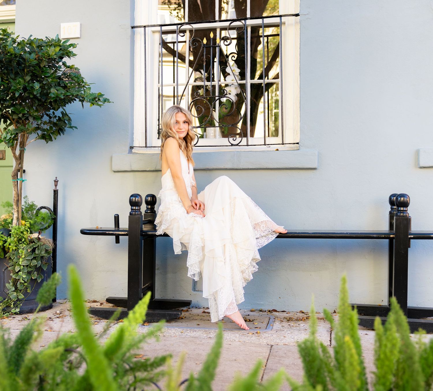 Bride in flowing white lace wedding dress sitting on black iron fence outside blue building surrounded by lush greenery.