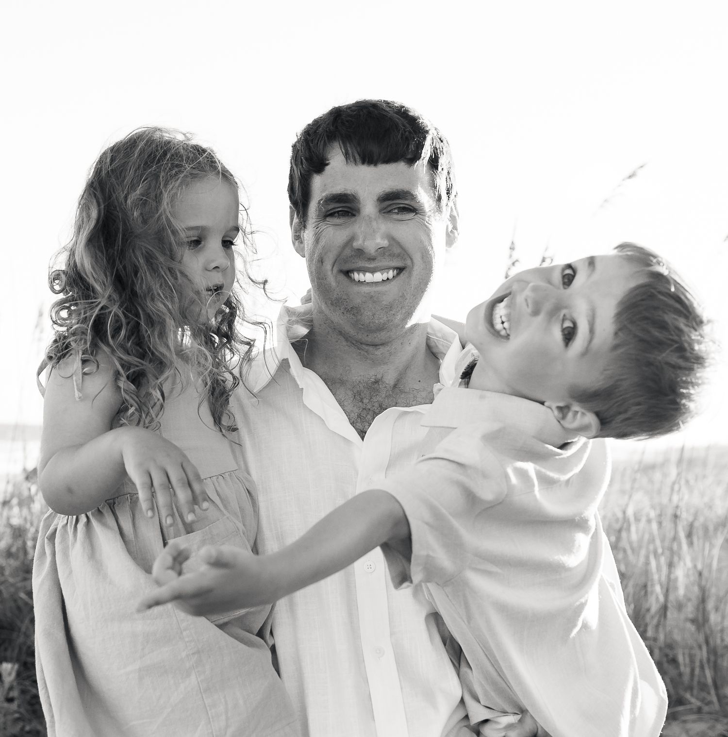 Black and white photo of a smiling man holding a young girl with curly hair and a laughing boy outdoors near tall grass.