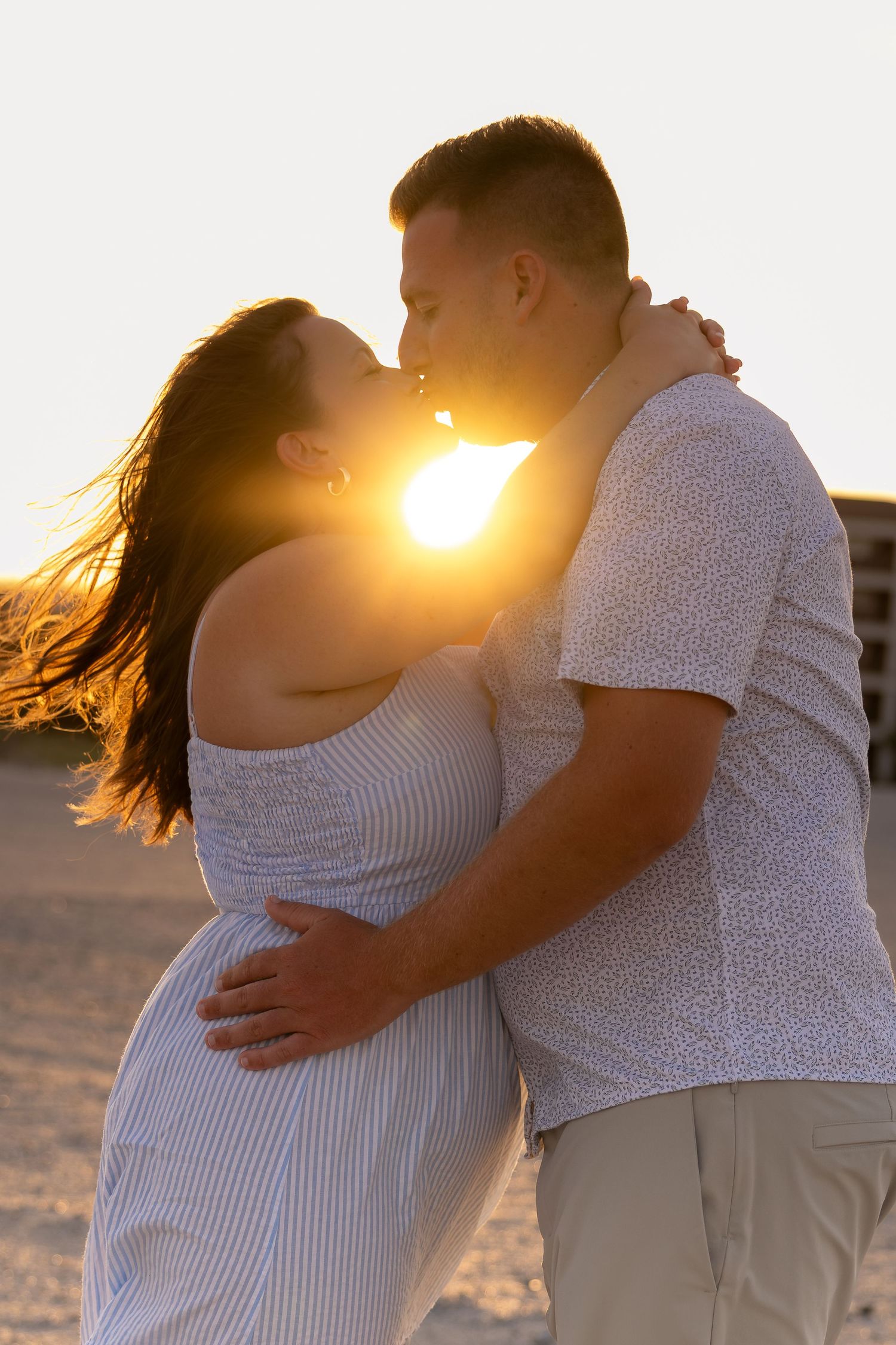 Couple sharing a kiss on the beach at sunset, with golden sunlight glowing between them and her long hair blowing in the breeze.