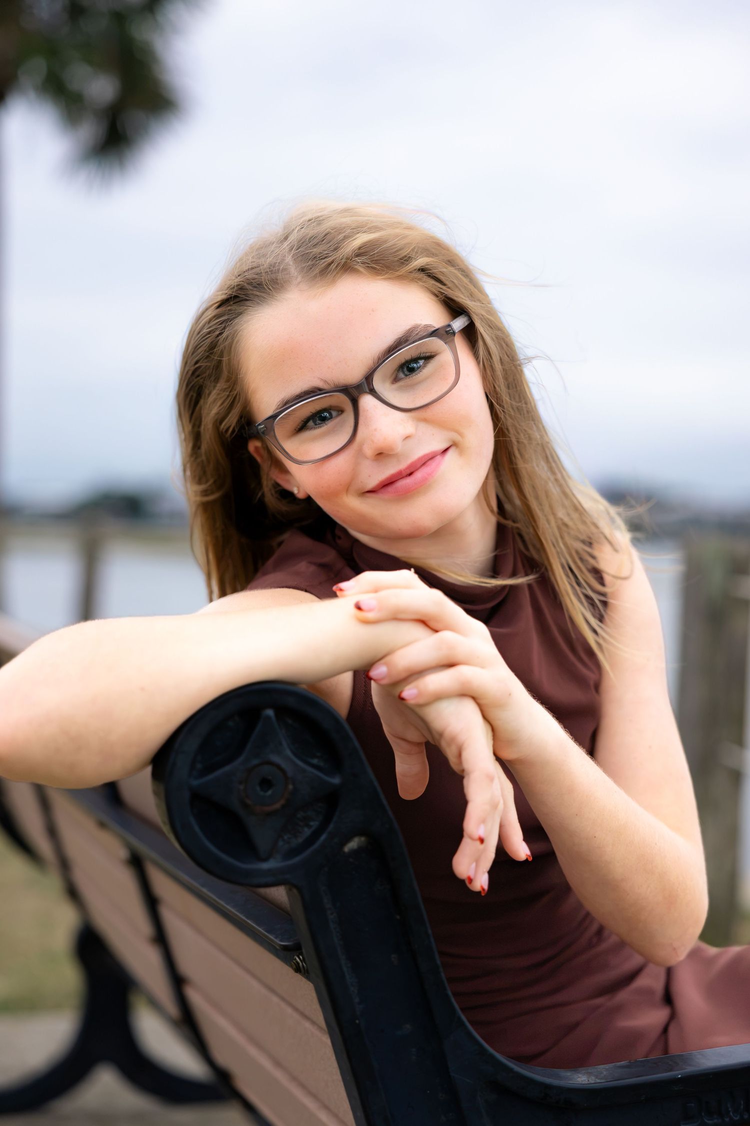 Smiling girl with glasses and long hair in a maroon sleeveless top leans on a black park bench with a waterfront blurred behind her.