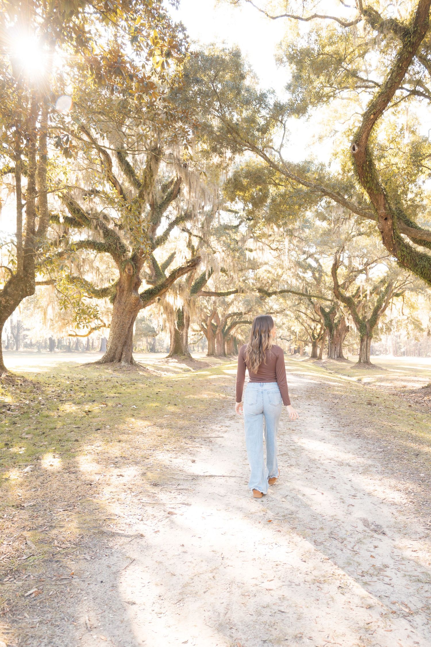 Woman walking down a sunlit path lined with mossy oak trees in a brown top and light blue wide-leg jeans.