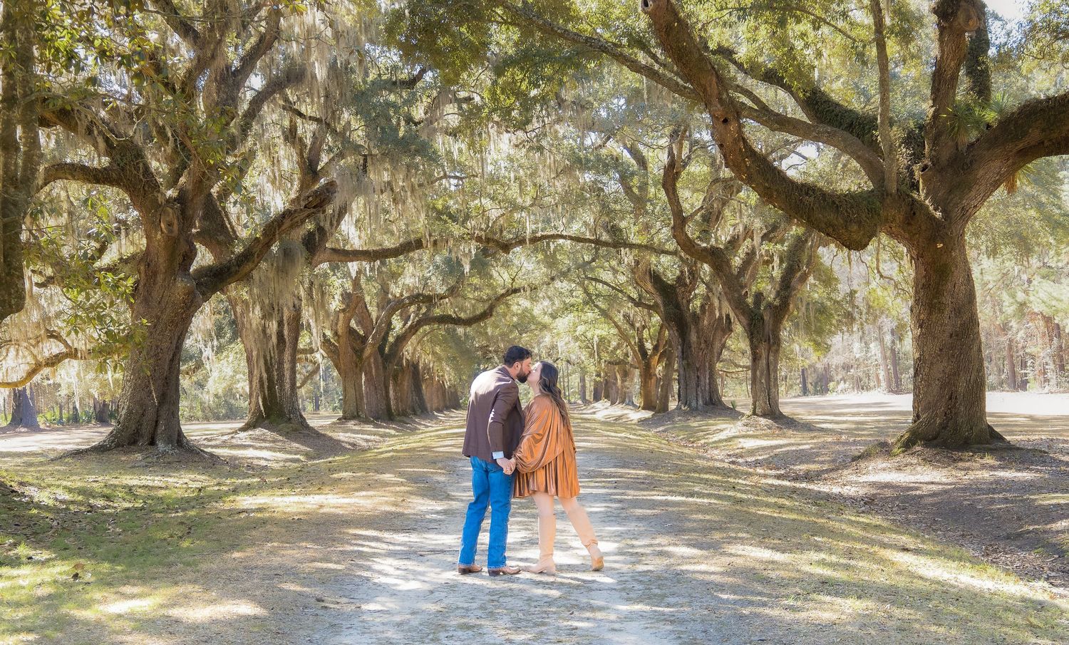 Couple sharing a romantic kiss under a canopy of moss-draped oak trees on a sunlit path.